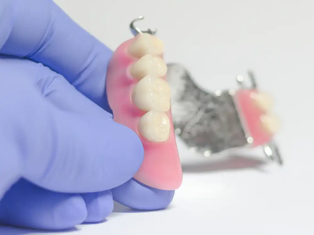 Gloved hand holding pink dental prosthetic with artificial teeth against white background