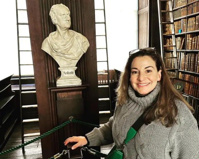 Woman smiling at camera in library with classical bust sculpture and bookshelves behind her.