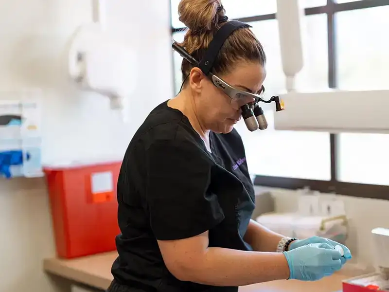 Woman in lab coat wearing protective eyewear examining sample in dental laboratory setting.