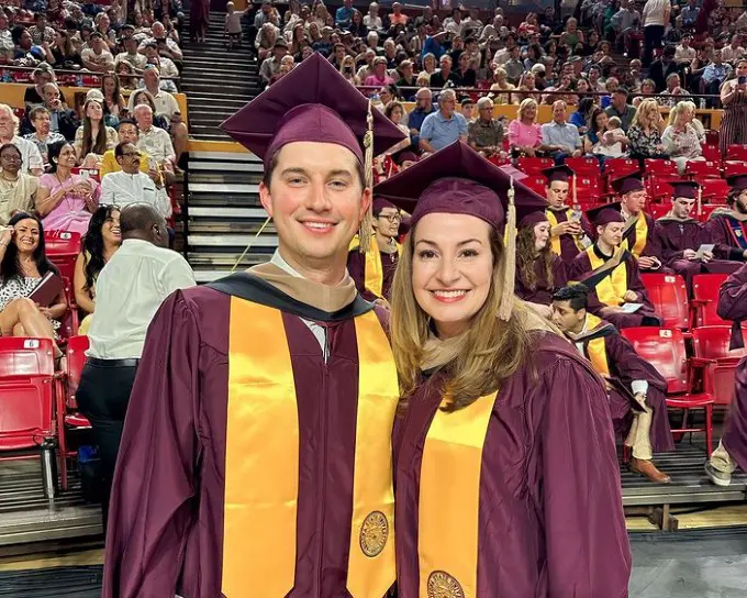 Two graduates in maroon and gold regalia smile at indoor graduation ceremony with crowded arena seating.