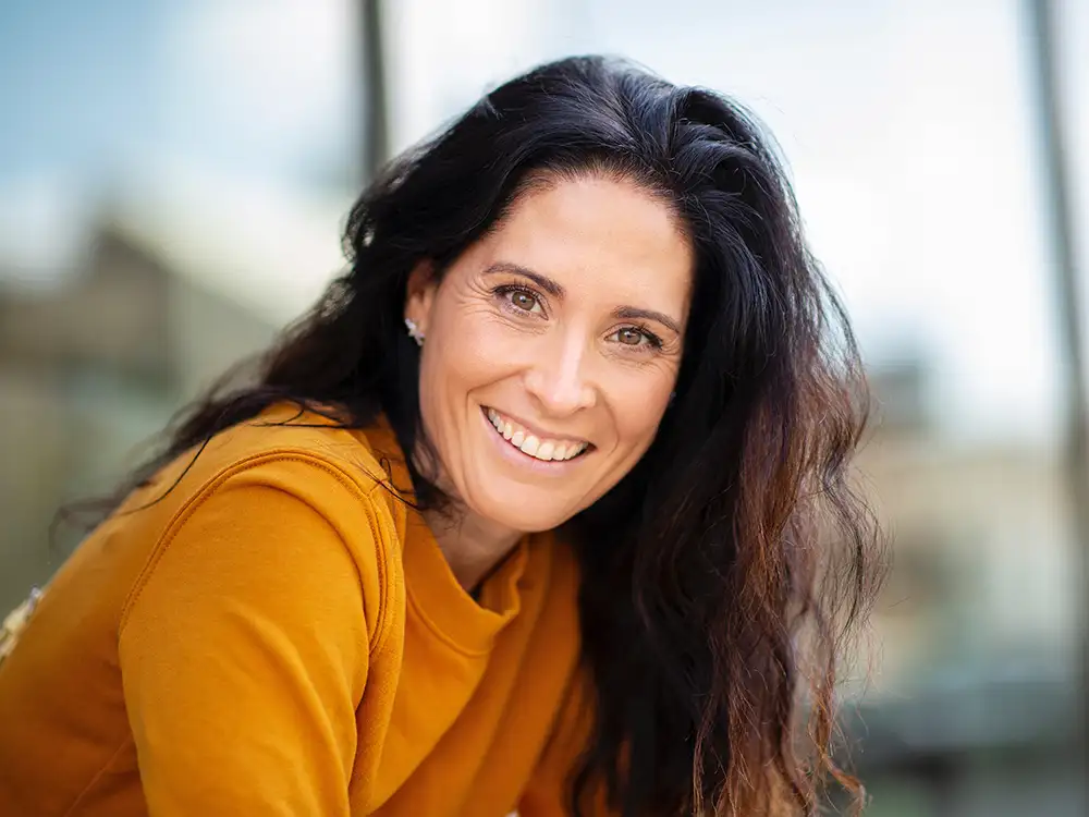 Woman in yellow shirt smiling outdoors with palm trees in blurred background.