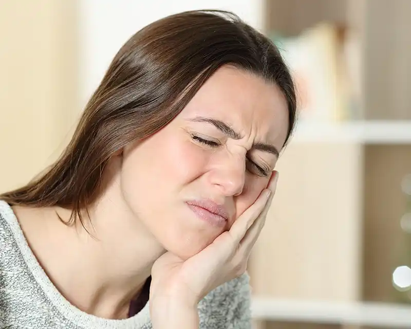 Woman with hand to cheek displaying discomfort or jaw pain indoors.