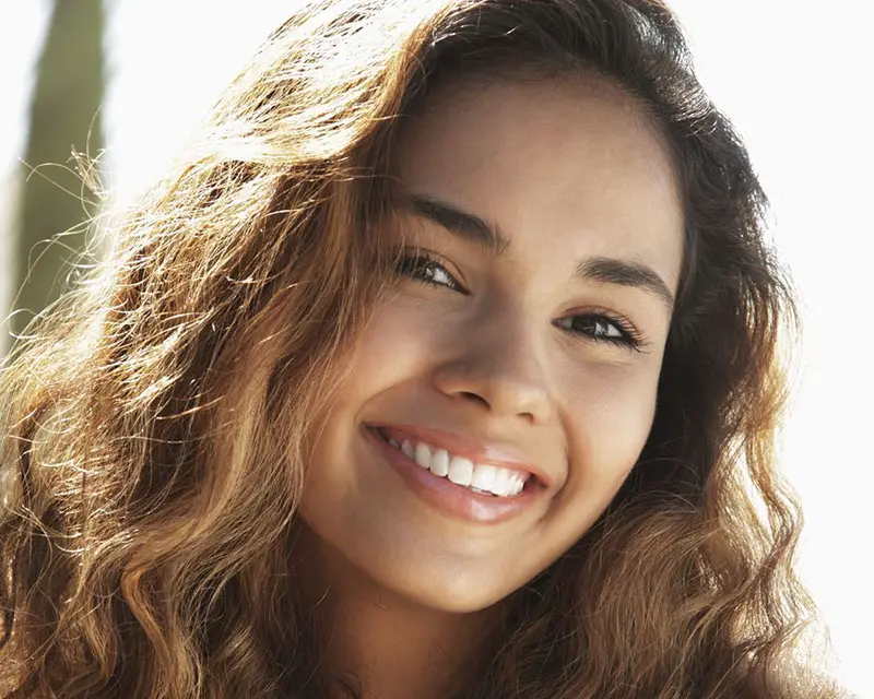 Young woman smiling at camera with wavy brown hair in natural sunlight